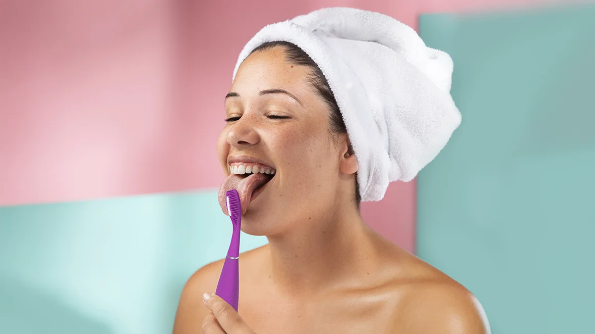 A young woman with her hair in a towel on a pink and mint background using issa 4 smile sonic toothbrush